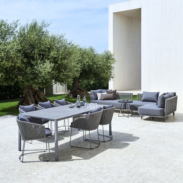 Relax and dine in style at this outdoor oasis, featuring a grey table, chairs, and a chic modular sofa, all nestled under the canopy of an olive tree.